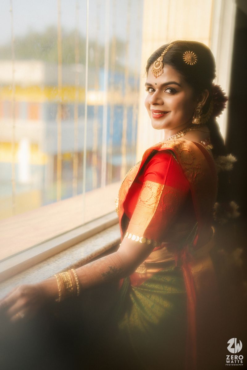 Bride in red and green silk saree with gold jewelry smiling by window.