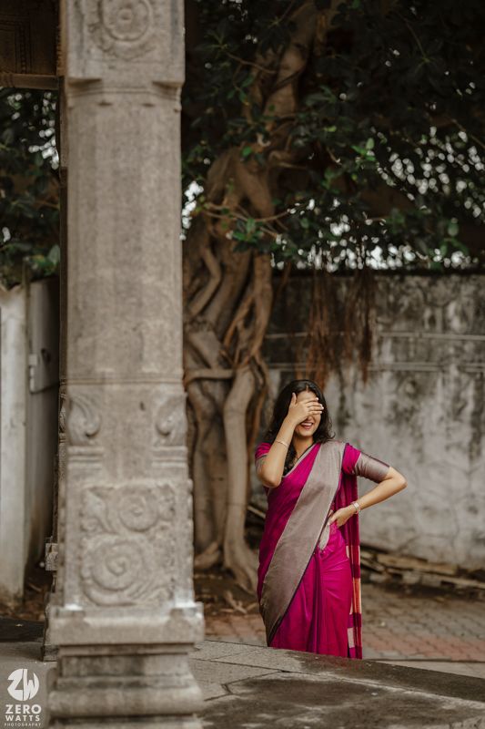 A woman in a pink sari poses playfully by an ornate stone pillar with trees in the background.