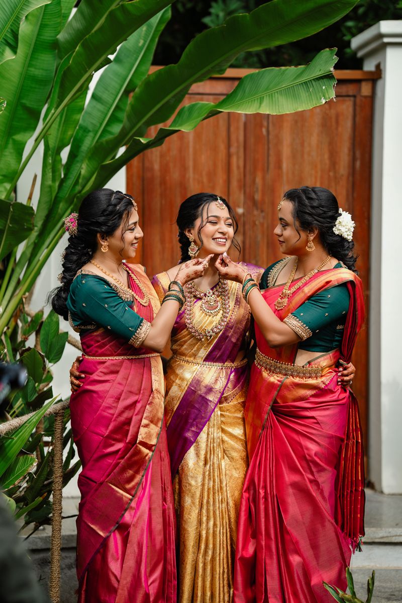 Women in vibrant silk saris share joyful moments together during a traditional celebration near tropical palm leaves.