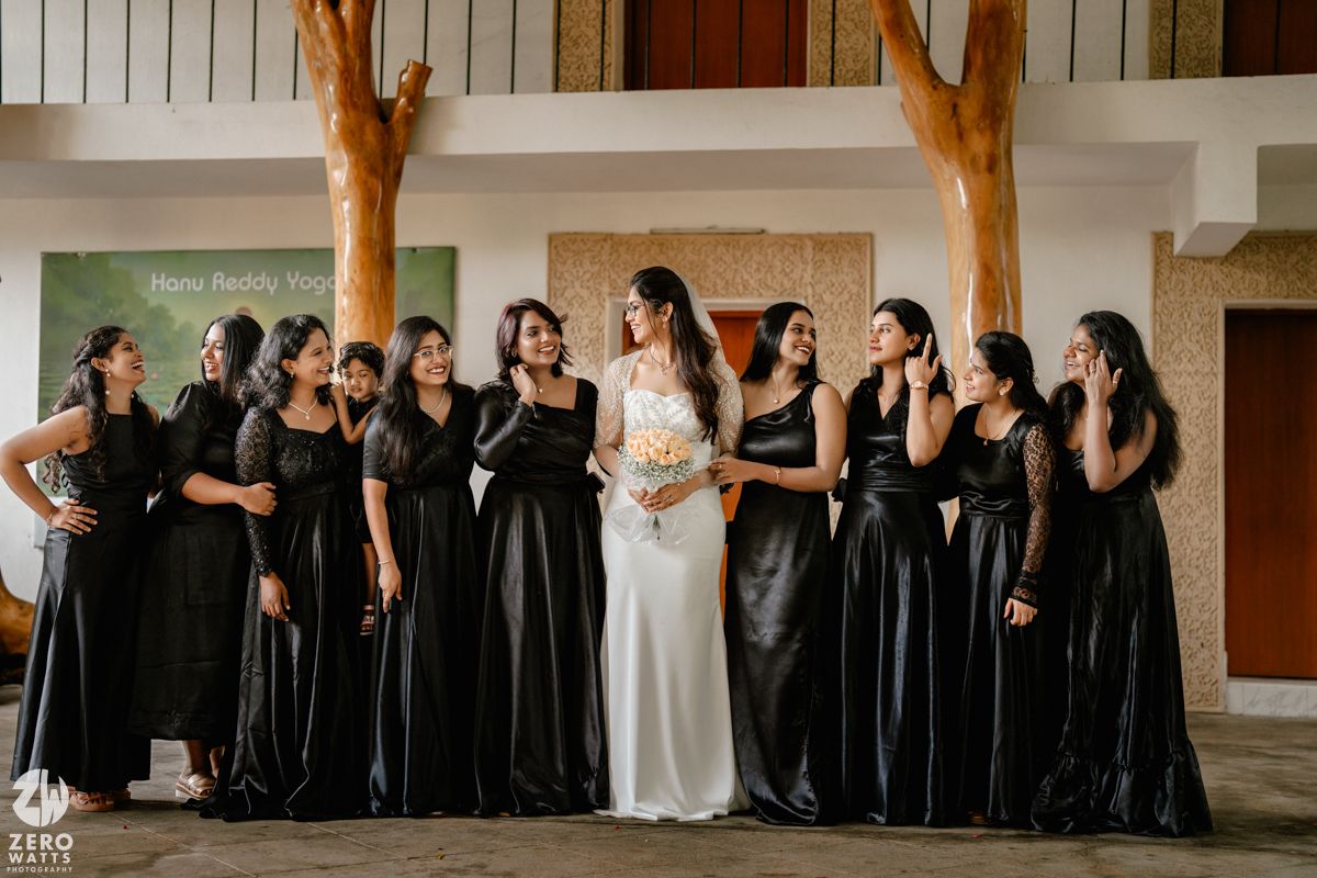 A bridal party poses together indoors wearing elegant white and black dresses in a rustic venue with wooden beams.