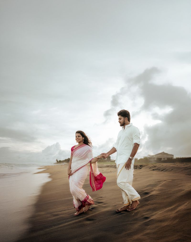Couple in traditional Indian attire runs joyfully across coastal sand dunes at sunset.