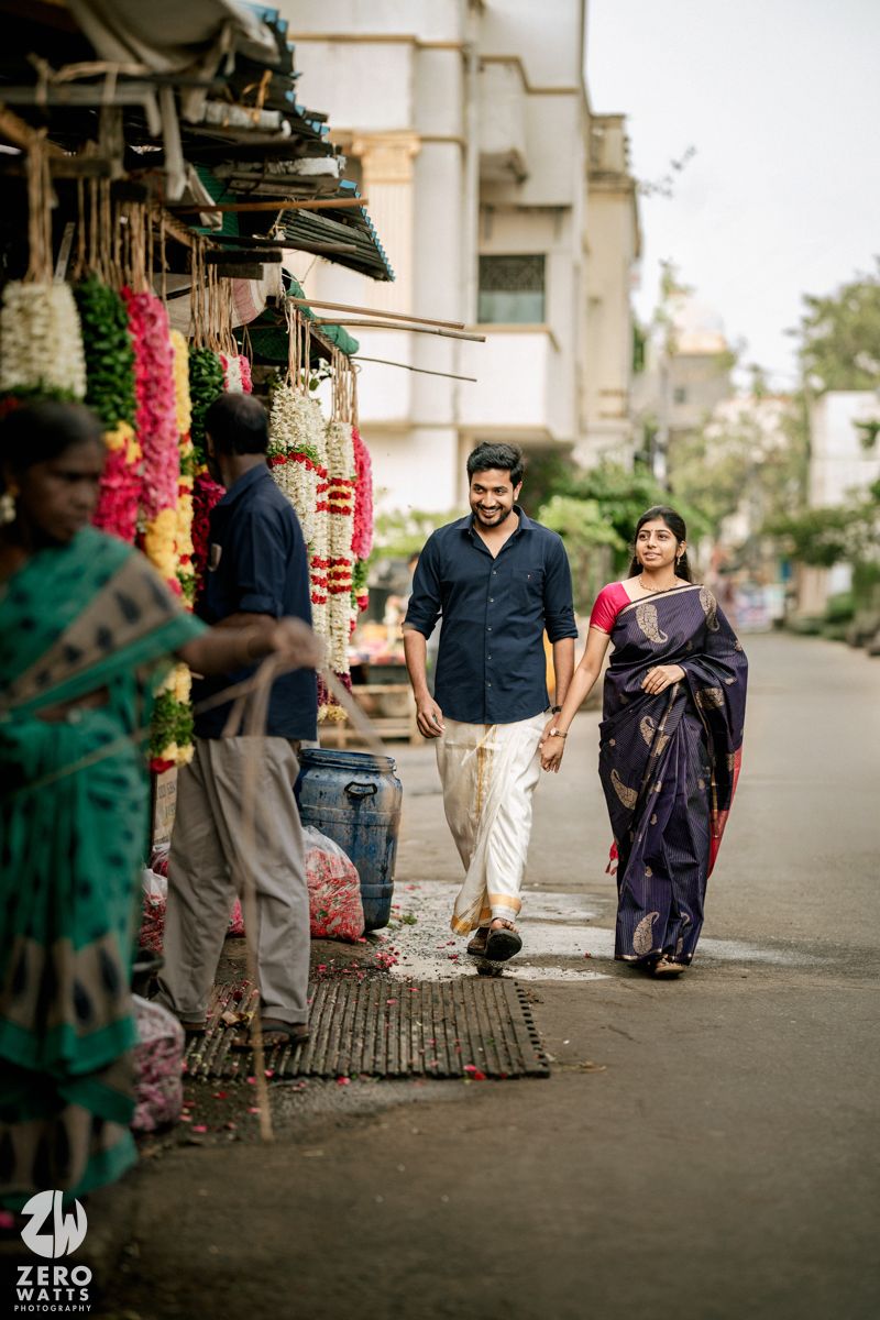 Couple walking and posing together near flower vendors on a street with buildings in the background.