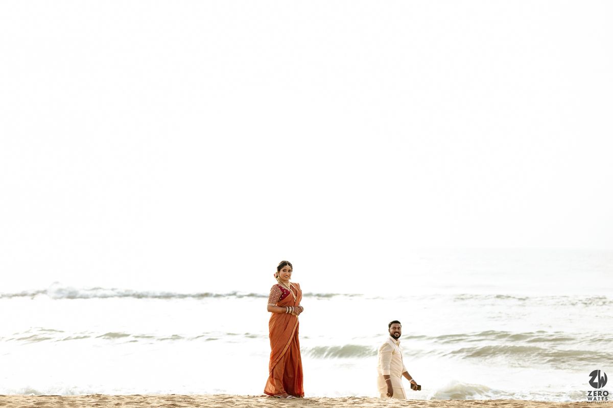 Two people walk along a sandy beach at sunset with waves crashing in the background.