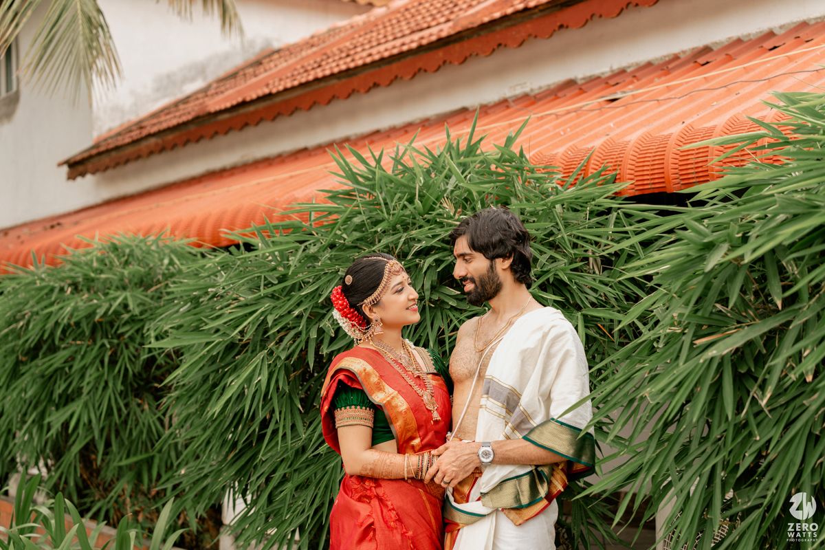 South Indian bride in red Kanjivaram saree and groom in white dhoti sharing a romantic moment outdoors.