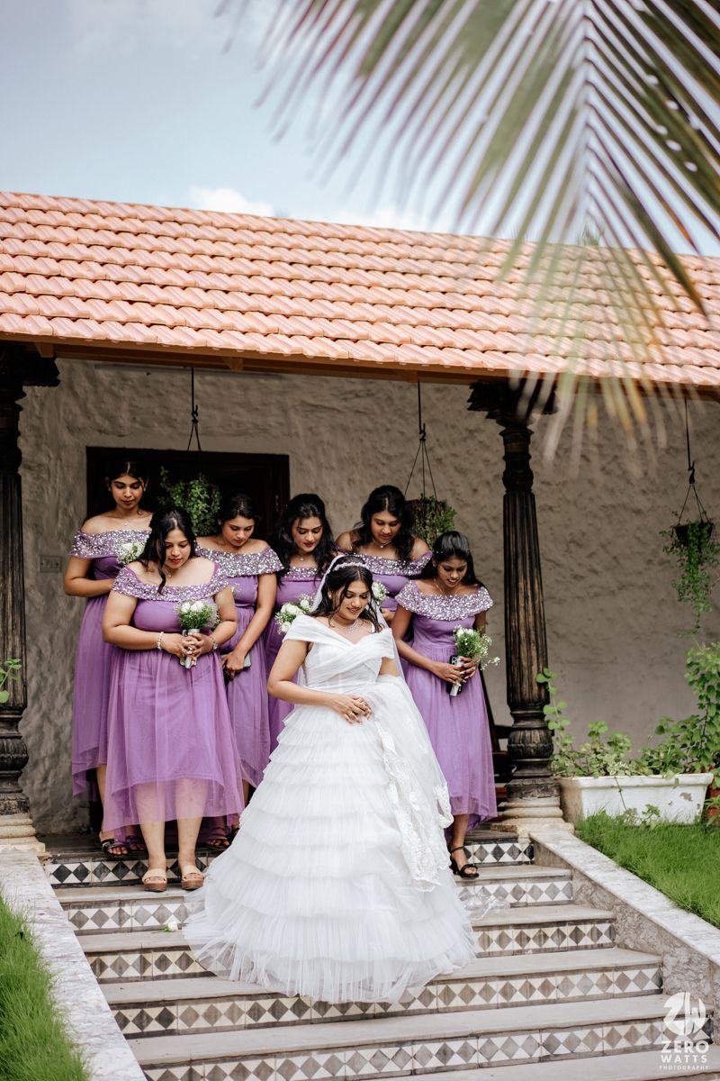 Bride in white gown poses with bridesmaids in lavender dresses on tiled steps outside a rustic venue.