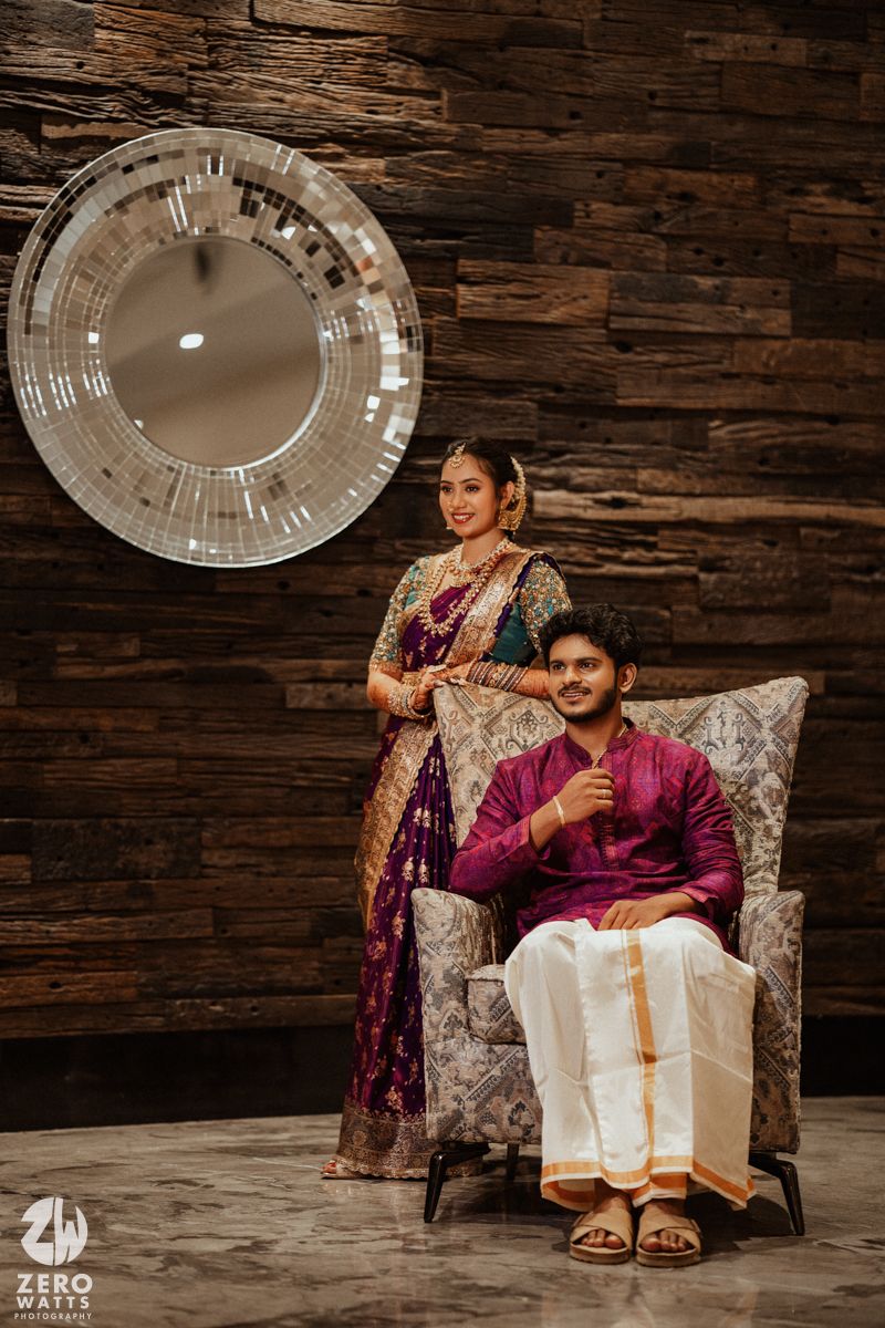 Indian couple in traditional attire pose elegantly, bride in purple saree, groom seated in chair.
