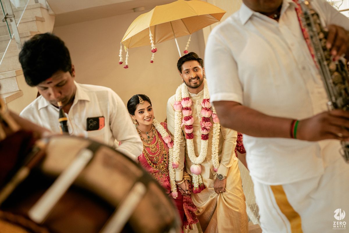 A bride and groom in traditional South Indian wedding attire with flower garlands stand under a yellow umbrella during their ceremony.