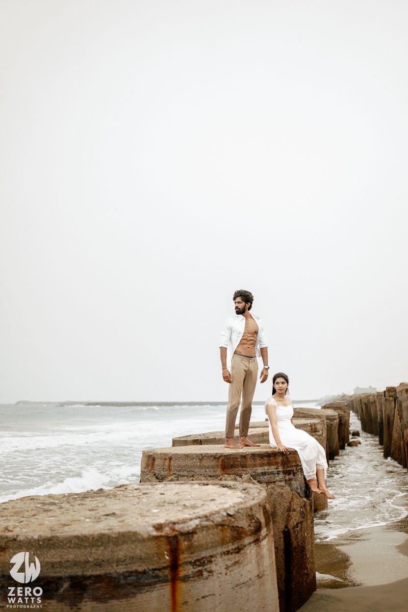 Couple posing on seaside stone pier, man in white shirt and beige pants standing, woman in white dress seated.