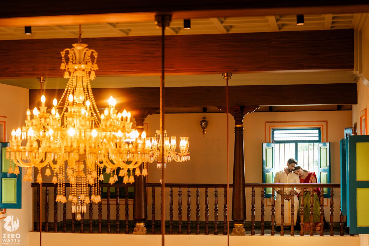 Couple sharing a romantic moment on a balcony beside an ornate golden chandelier in a heritage venue.