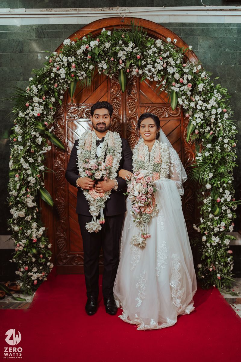 A couple stands under an elegant floral arch with white flowers and greenery against ornate wooden doors.