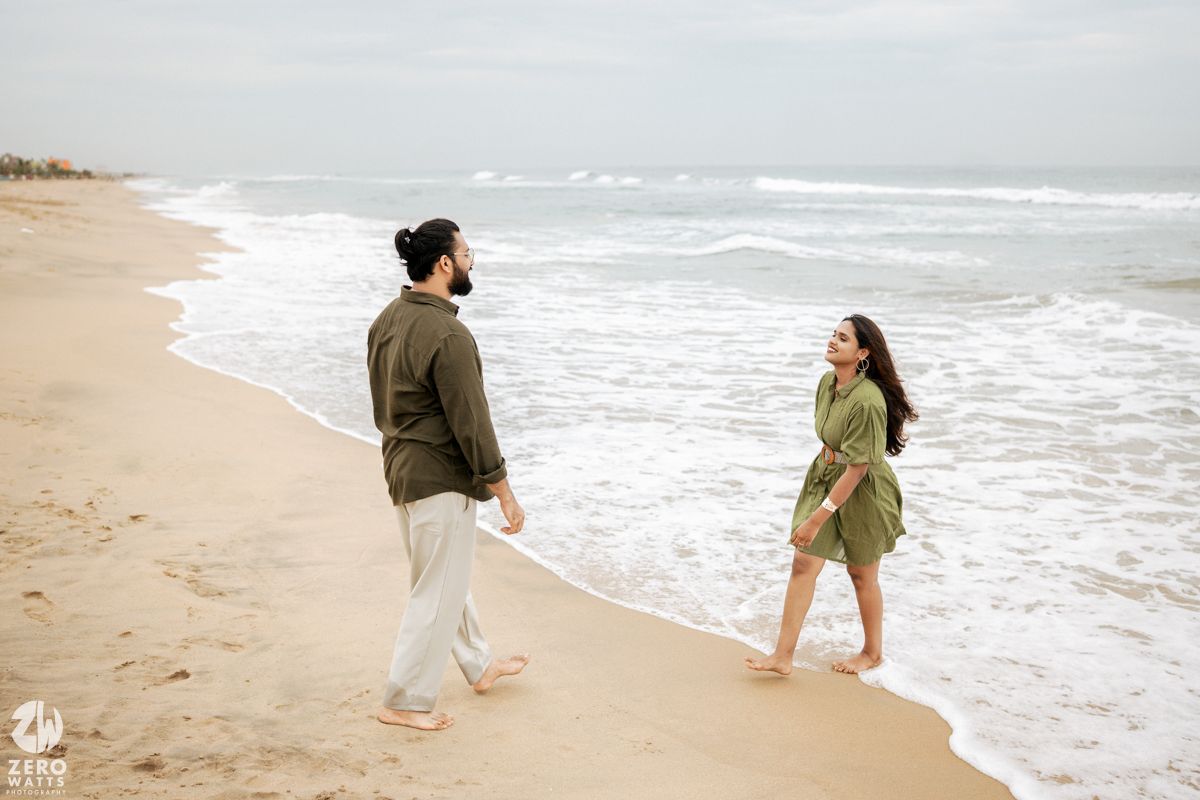 A couple walks along a misty beach shoreline with waves crashing against the sand on an overcast day.