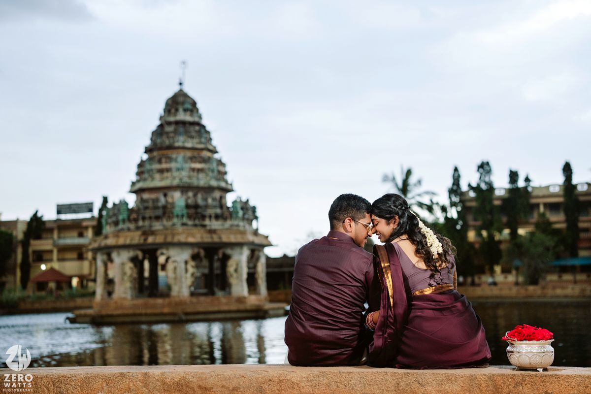 Romantic couple in burgundy outfits posing near traditional Indian temple architecture at sunset.