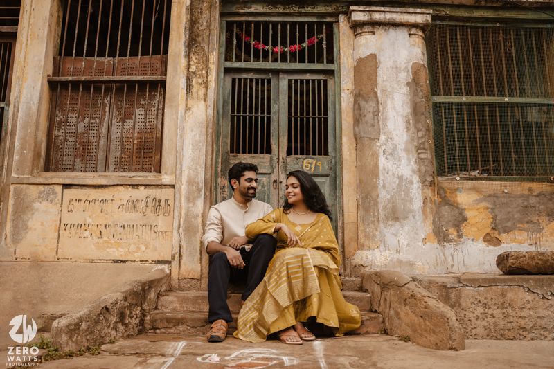 A couple sits together on weathered stone steps against an old colonial building facade.