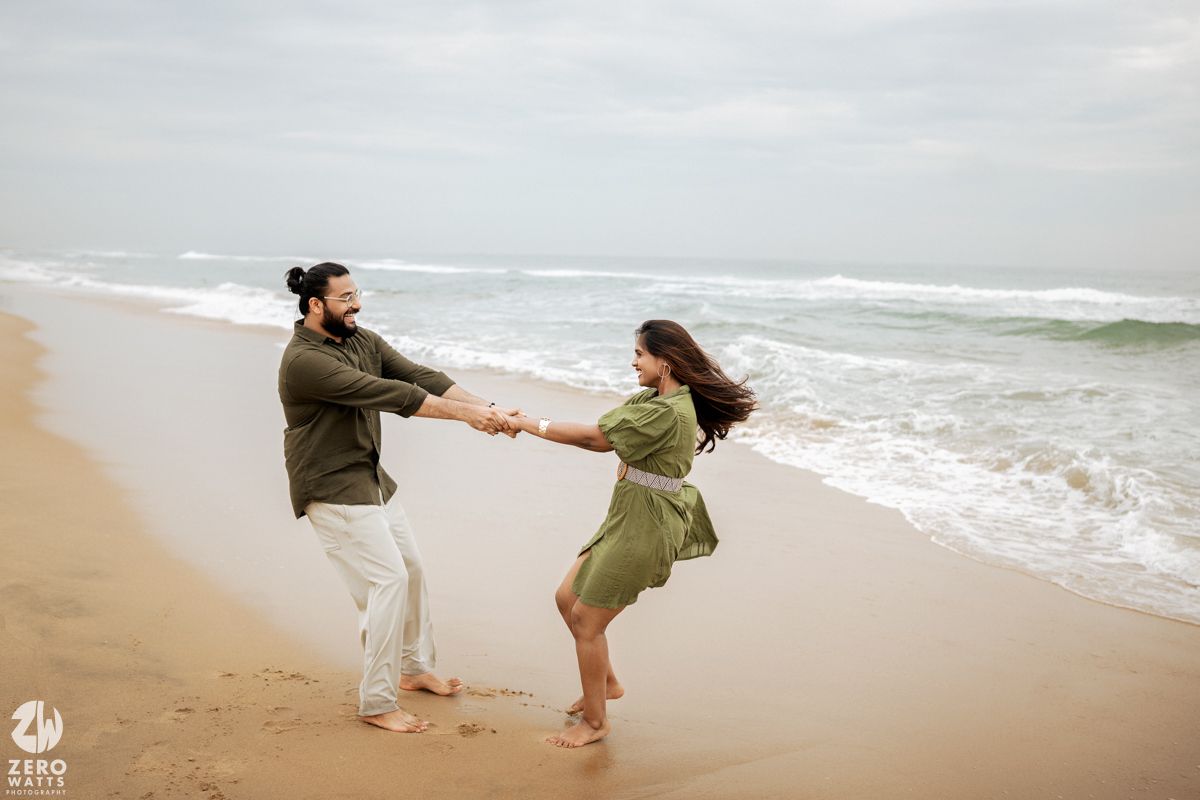 A couple in green outfits holds hands and spins playfully on a cloudy beach by the ocean waves.