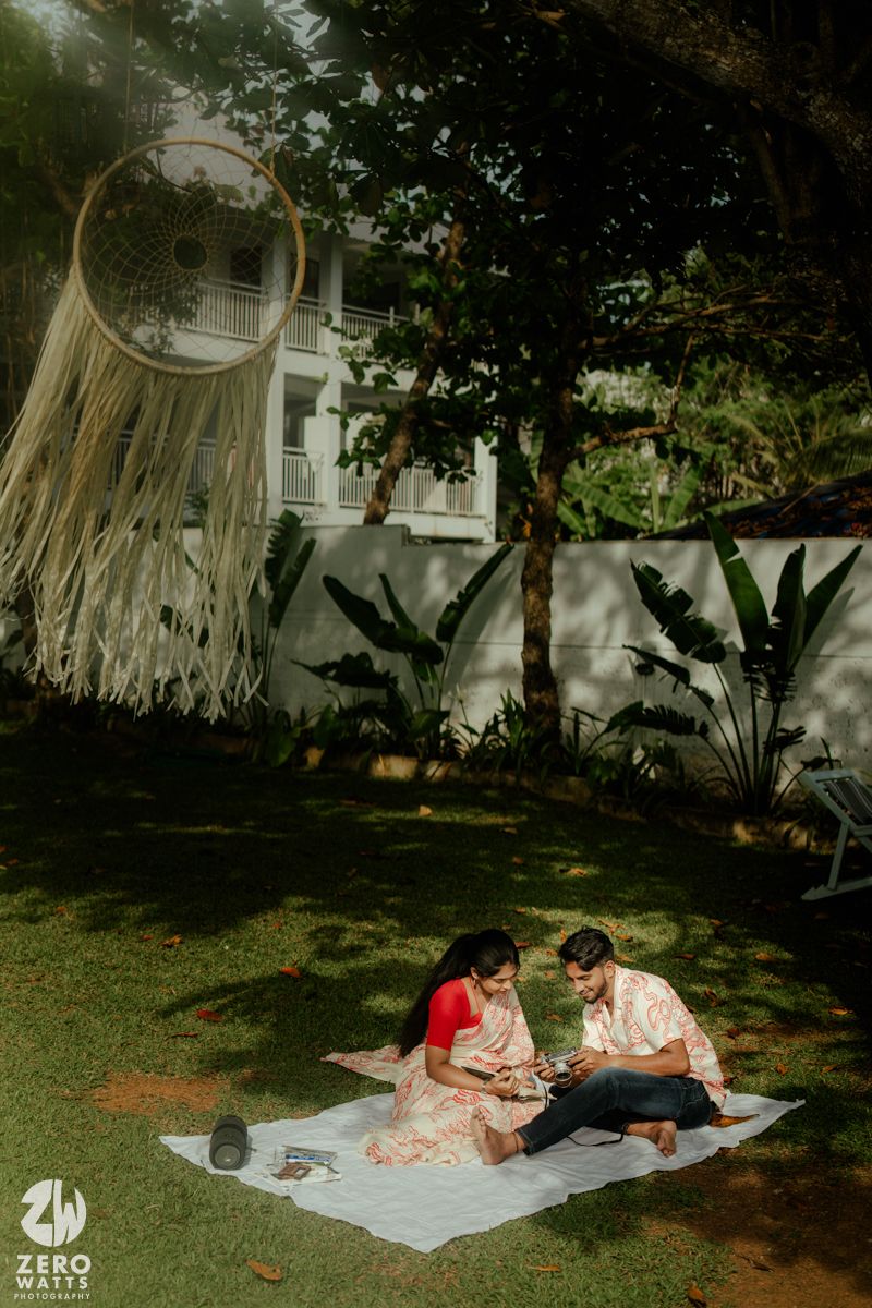 A couple relaxes on a picnic blanket under the shade of large trees in a garden setting.