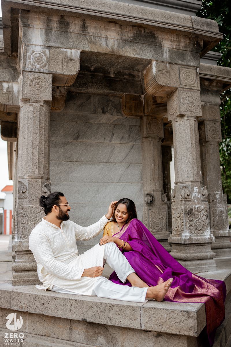 Two people in contrasting white and purple outfits lounge on a decorative stone balcony in a romantic pose.