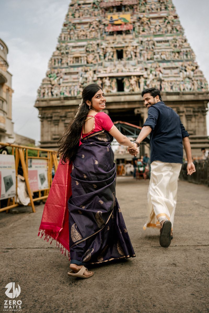 A couple in traditional Indian attire dances playfully in front of an ornate temple with intricate architectural details.
