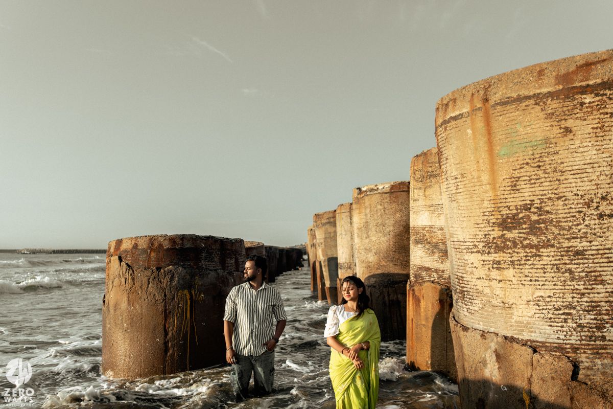 A romantic couple poses against stone pillars at the beach during golden hour.