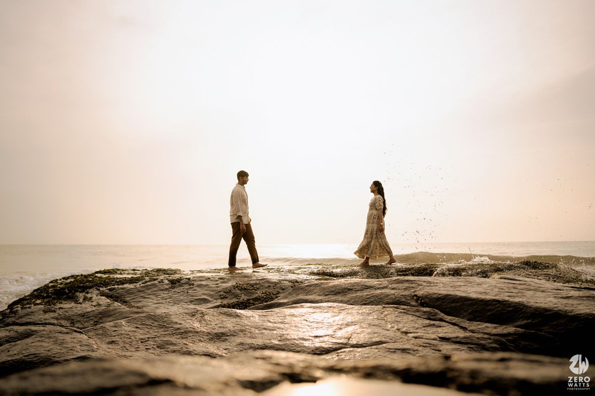 Silhouettes of two people standing on rocky shore during golden hour with dreamy coastal backdrop.