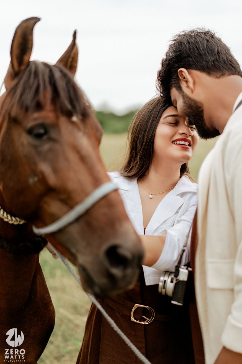 A couple shares intimate moments alongside a brown horse in a grassy field.