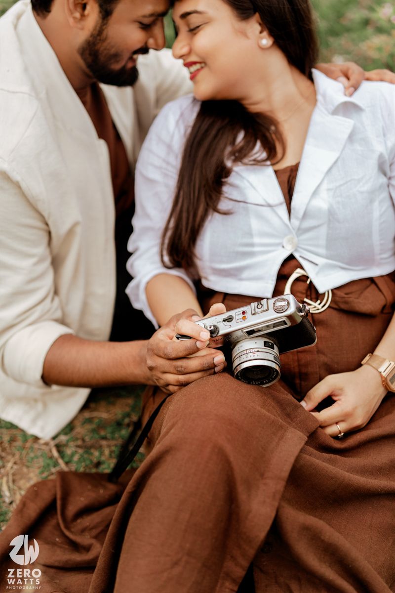 A romantic couple in white and brown outfits share tender moments while holding a vintage camera outdoors.