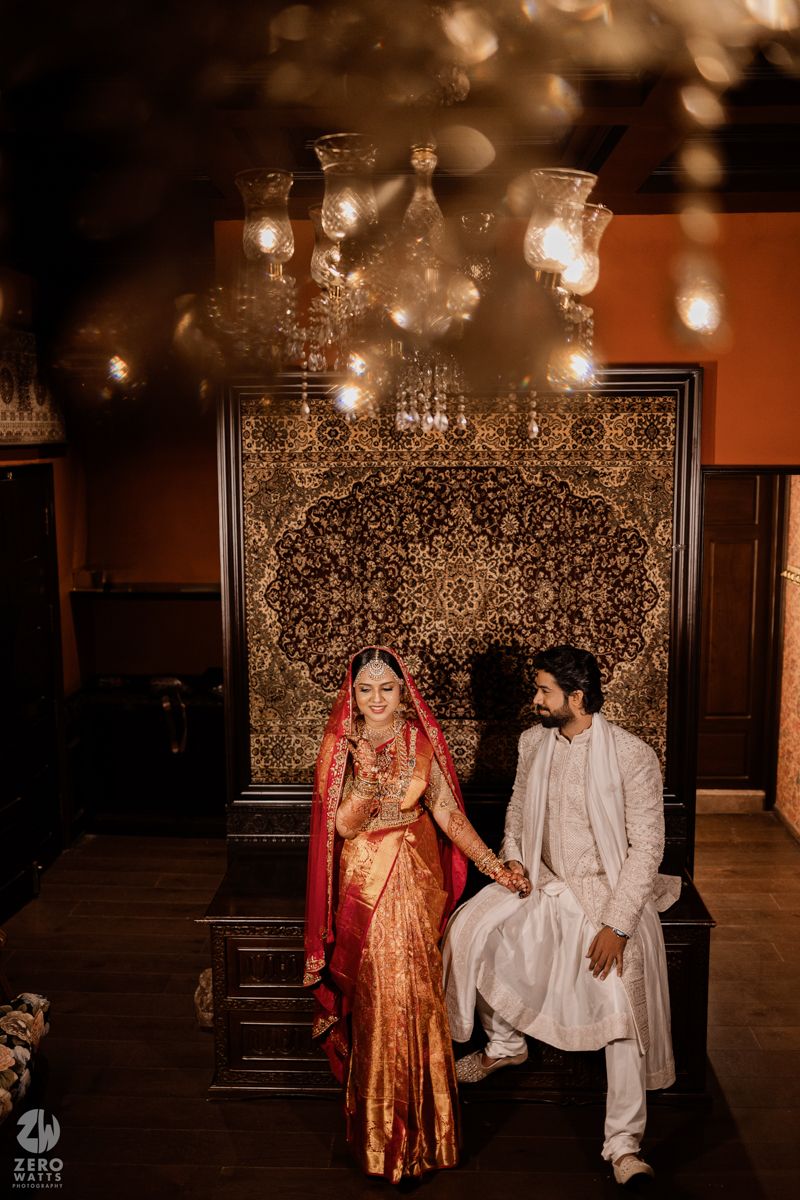 Couple in traditional wedding attire seated on ornate throne against illuminated decorative backdrop.
