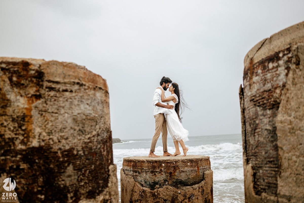 Couple embracing on rocky seaside formation with ocean waves, wearing white wedding attire in a romantic pre-wedding shoot.