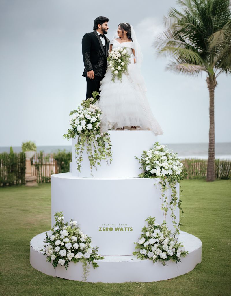 Couple in wedding attire stand atop a white tiered cake-shaped floral display at a beachside venue.