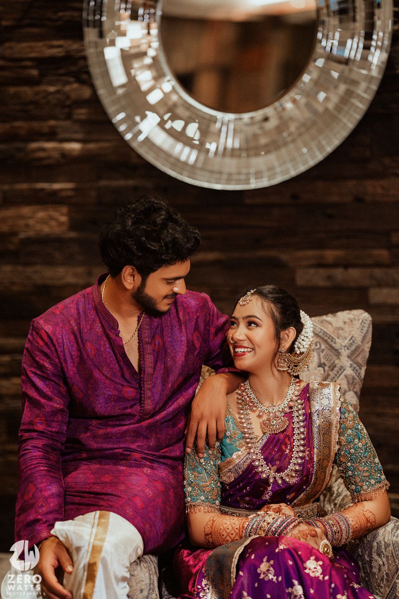 Couple in matching purple Indian wedding attire sitting together, bride adorned with traditional jewelry, smiling joyfully.