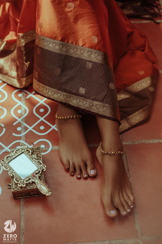 Close-up of Indian bride's feet adorned with gold anklets beside an ornate mirror on a traditional kolam floor.