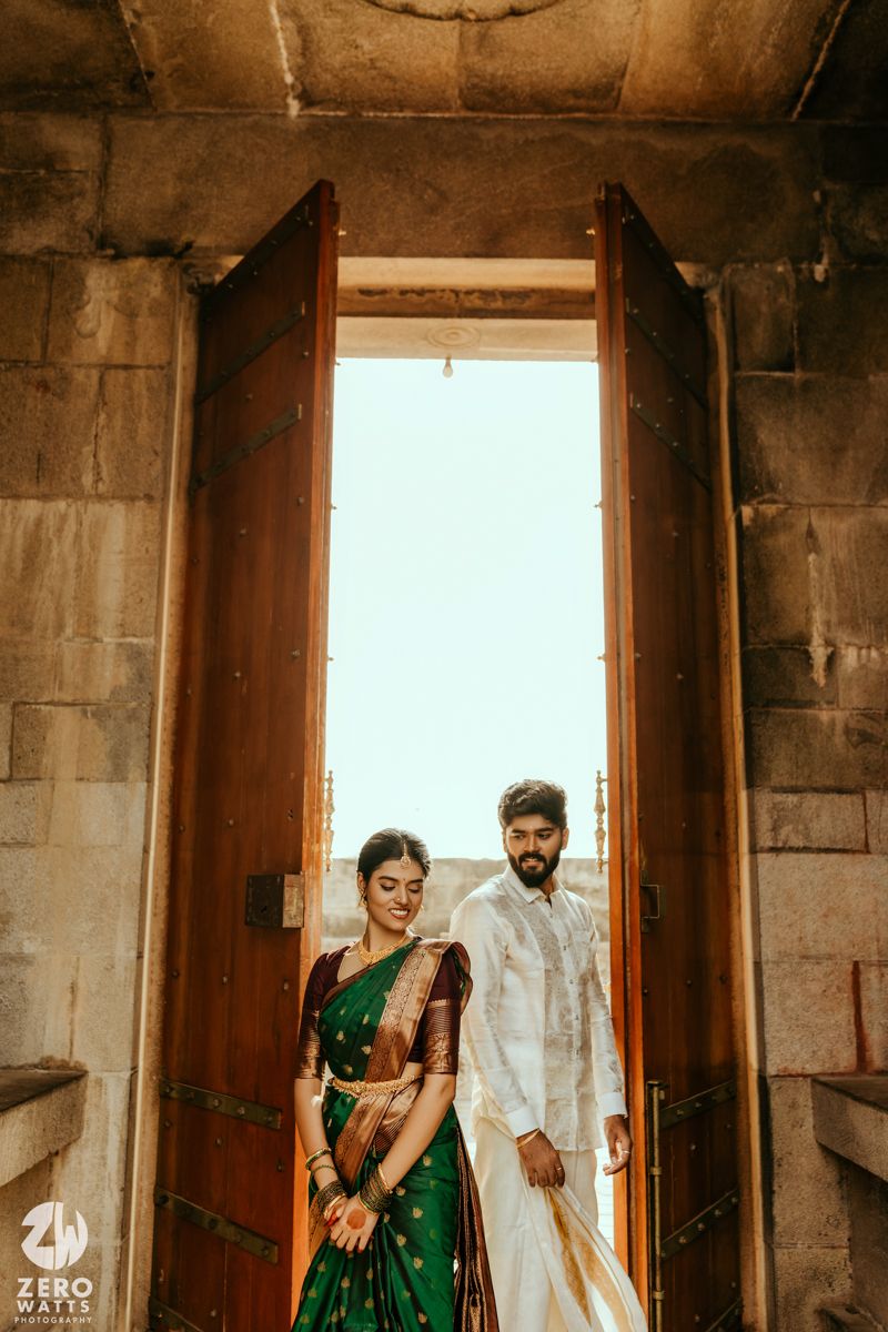 Indian bride in green Kanjivaram saree and groom in white sherwani standing at ornate temple doorway.
