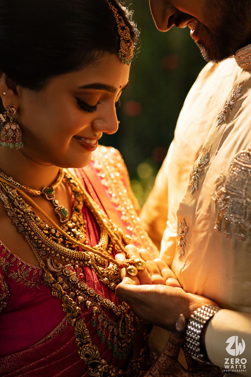 Bride adorned in gold jewelry and pink saree shares intimate moment with groom at Indian wedding.