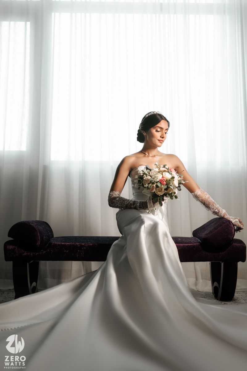 Bride in strapless white gown with long train holds bouquet, seated by sheer curtains in elegant bridal portrait.