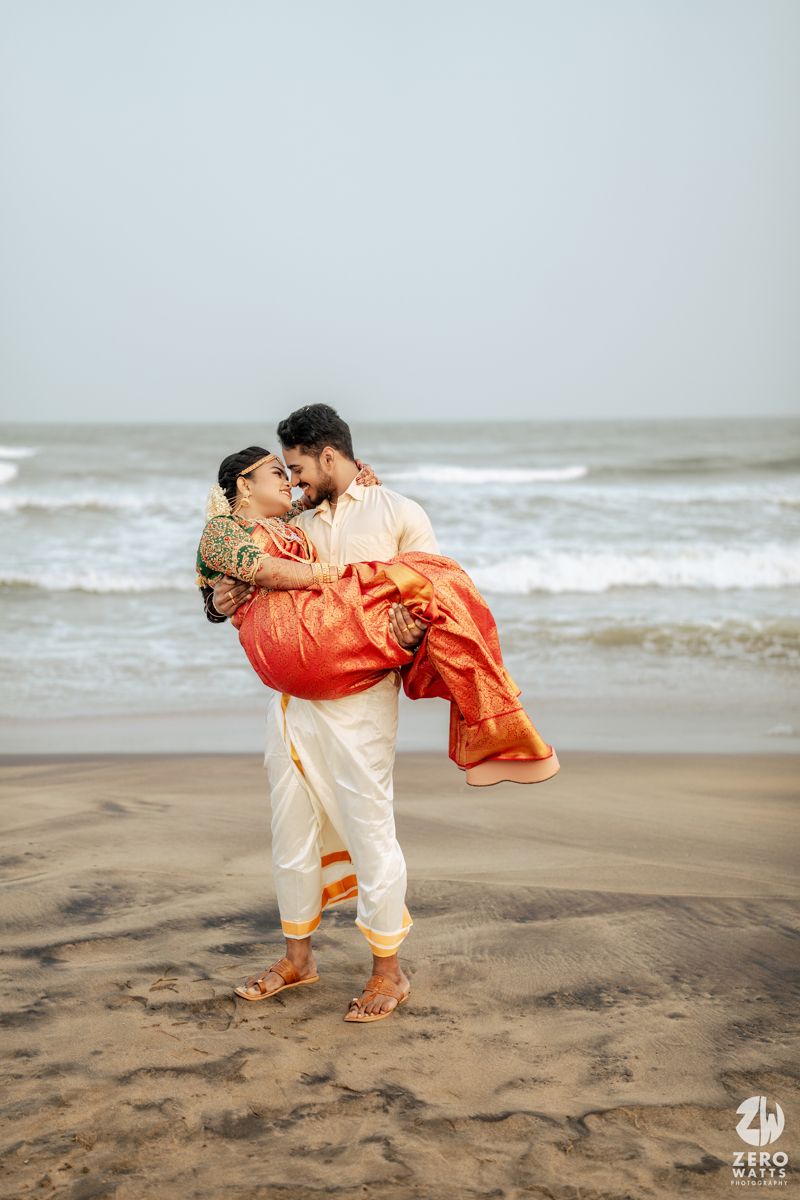 Indian groom carries bride in orange Kanjivaram saree on sandy beach with ocean waves in background.