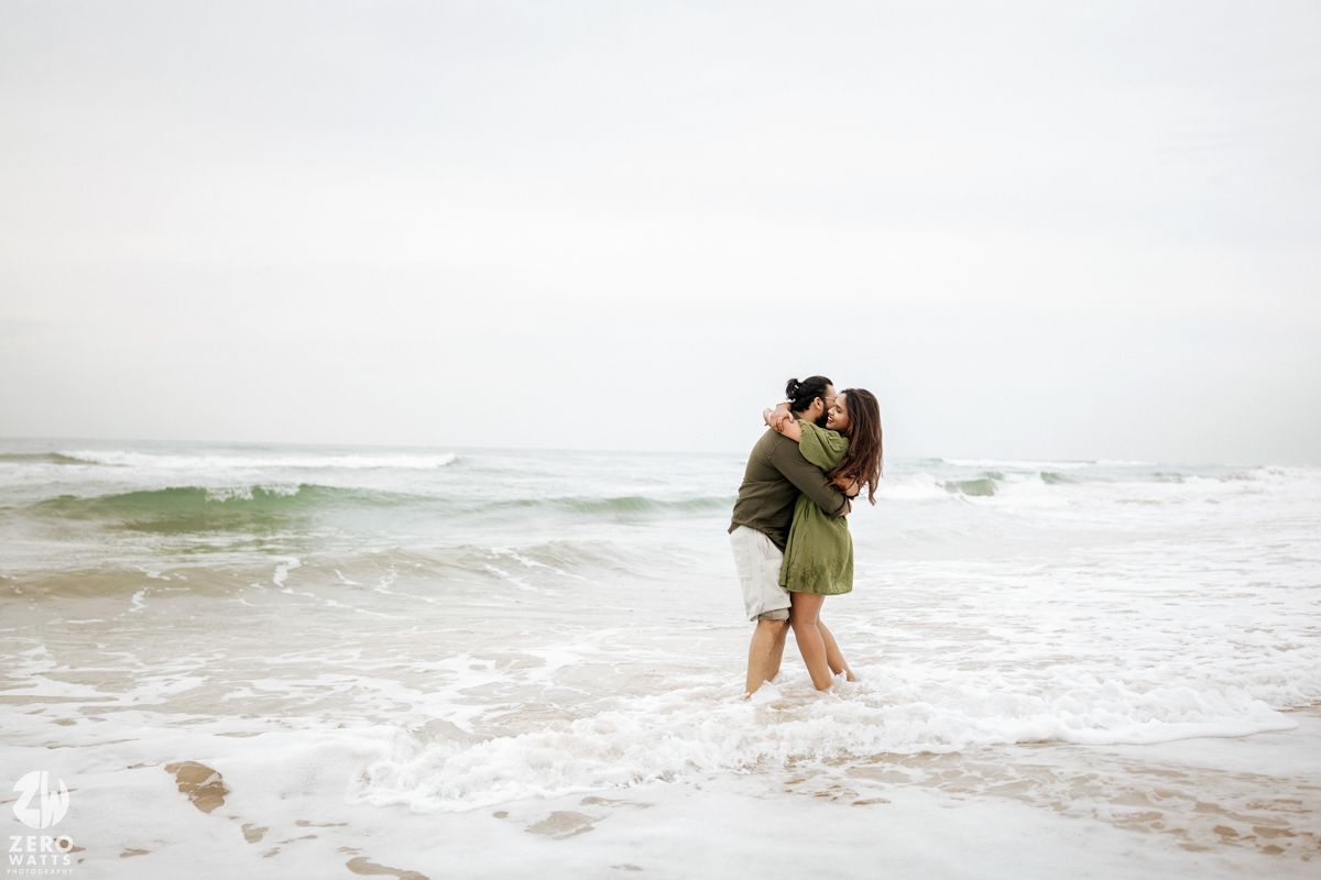 A couple embraces while standing in shallow ocean waves on a misty beach.