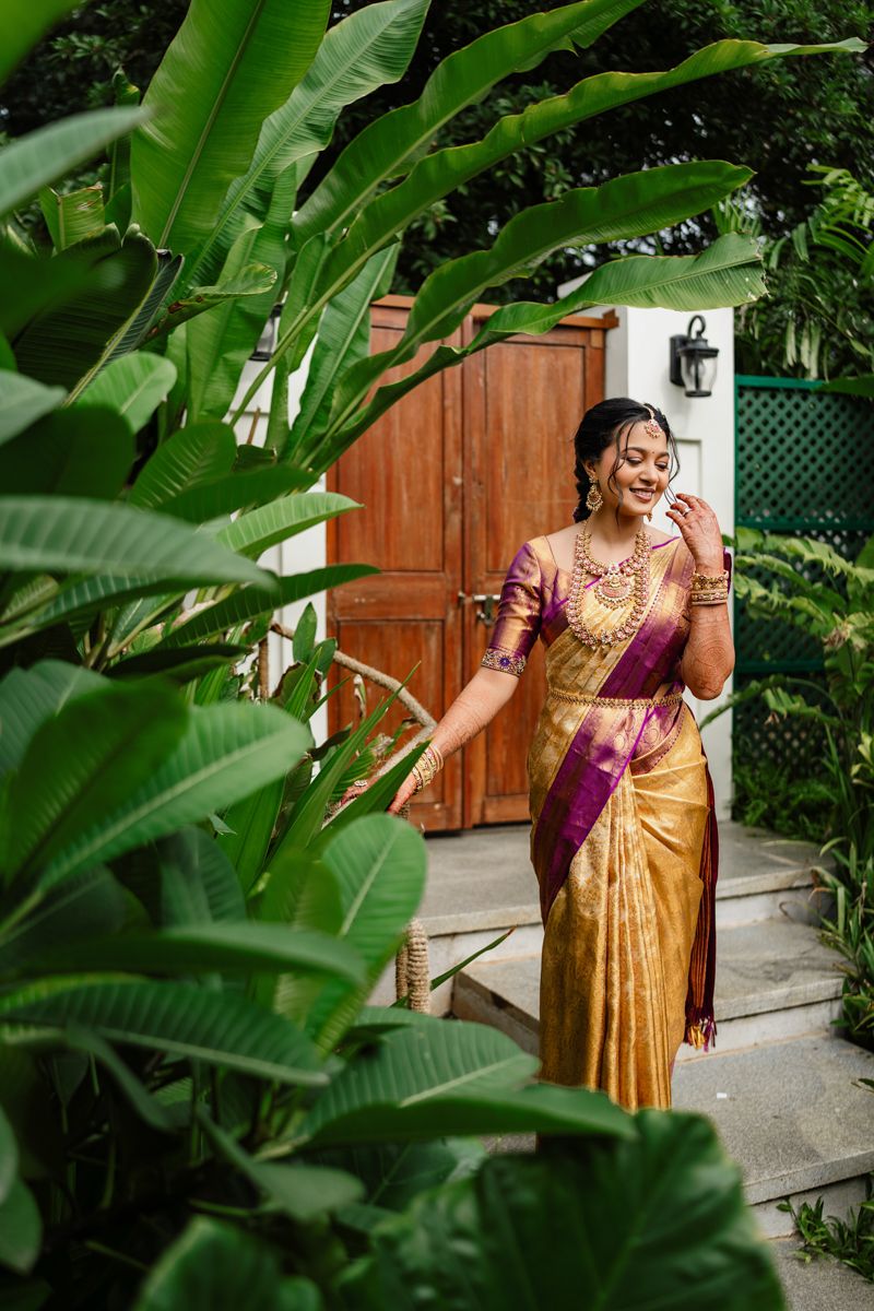 A person in a golden silk sari poses elegantly among lush green tropical foliage in a garden setting.