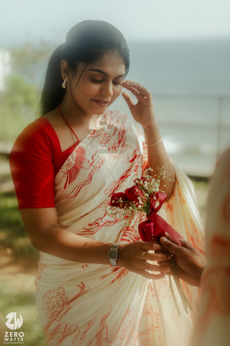 A woman in a white and red saree holds a small bouquet of roses near the ocean.