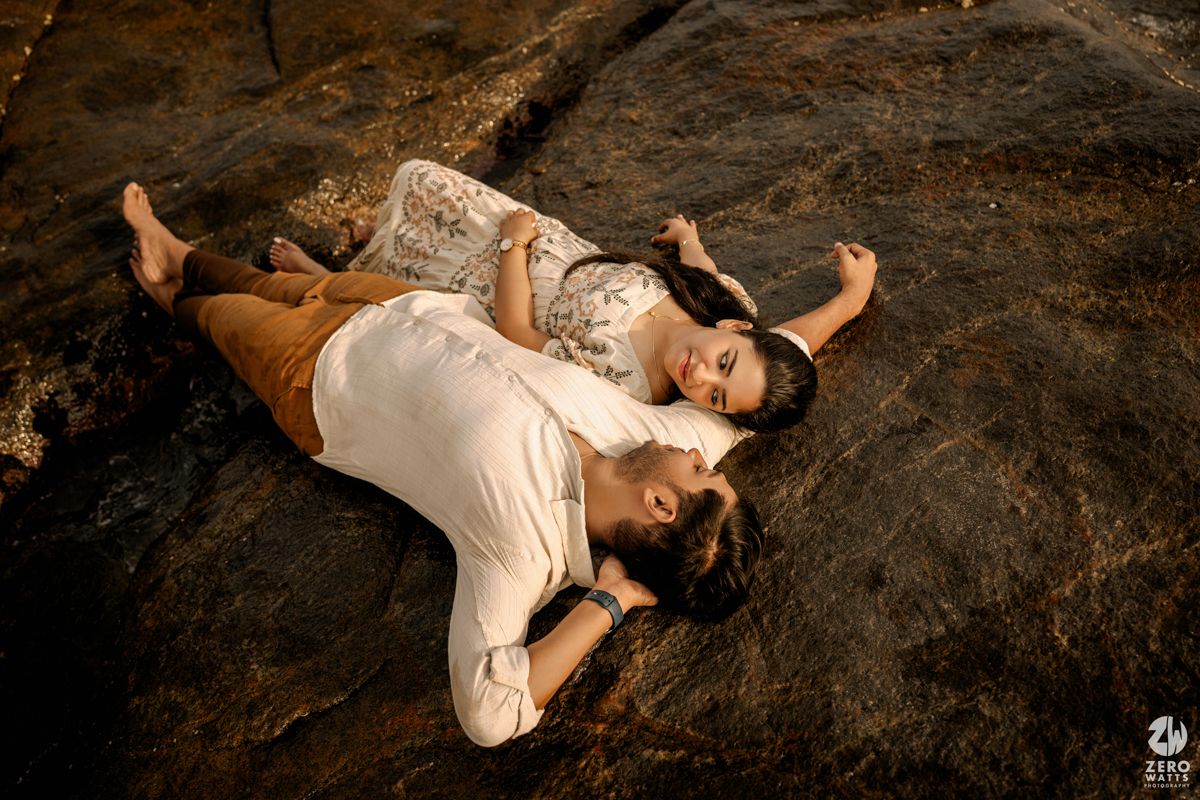Artistic couple's pose on dark rocky shore during dramatic sunset photoshoot.