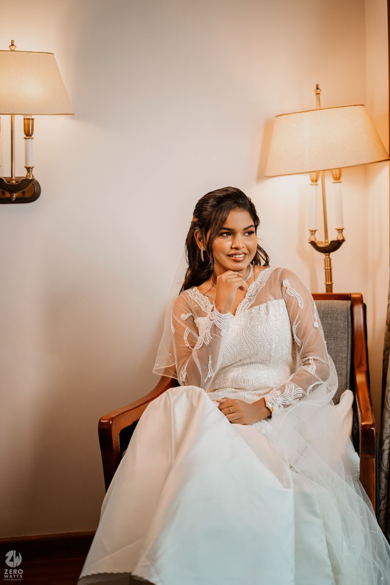 Bride in white lace gown sits elegantly in chair with soft lamp lighting creating romantic ambiance.