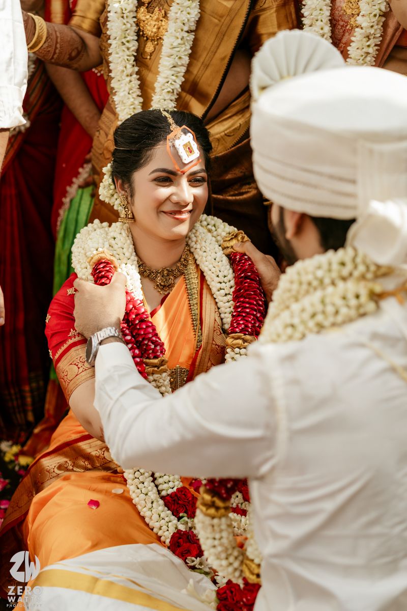 Bride in orange saree and red blouse smiles during traditional South Indian wedding garland exchange ritual.