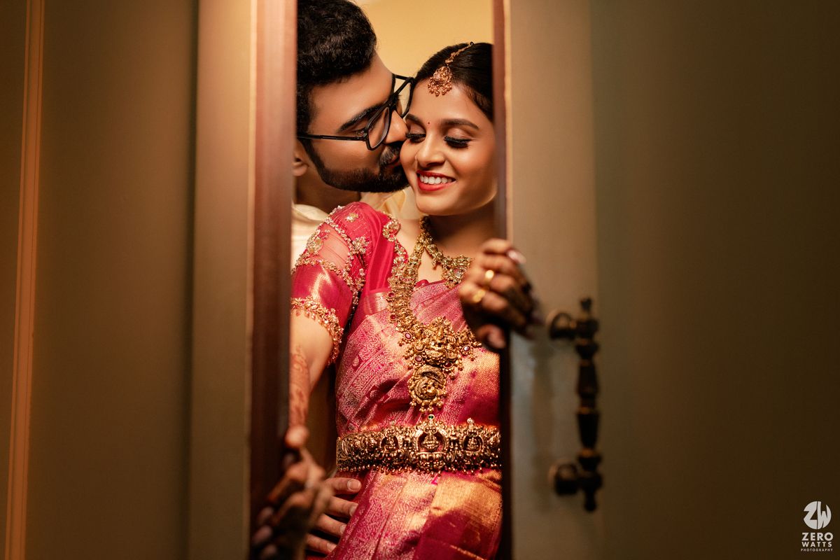 South Indian couple in traditional wedding attire share a romantic moment peeking through a doorway.