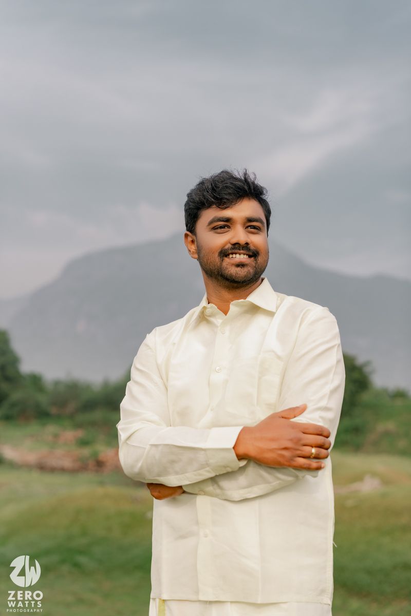 A smiling Indian man in a white kurta stands with arms crossed against a scenic mountain backdrop.