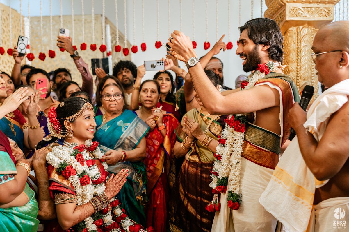 Traditional South Indian wedding ceremony with bride in silk saree and groom exchanging garlands surrounded by joyful family.
