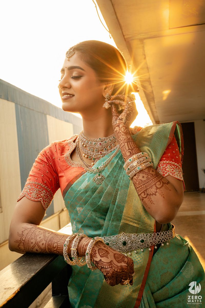 Indian bride in teal saree with coral blouse, adorned with mehndi, jewelry, and bangles, posing in golden sunlight.