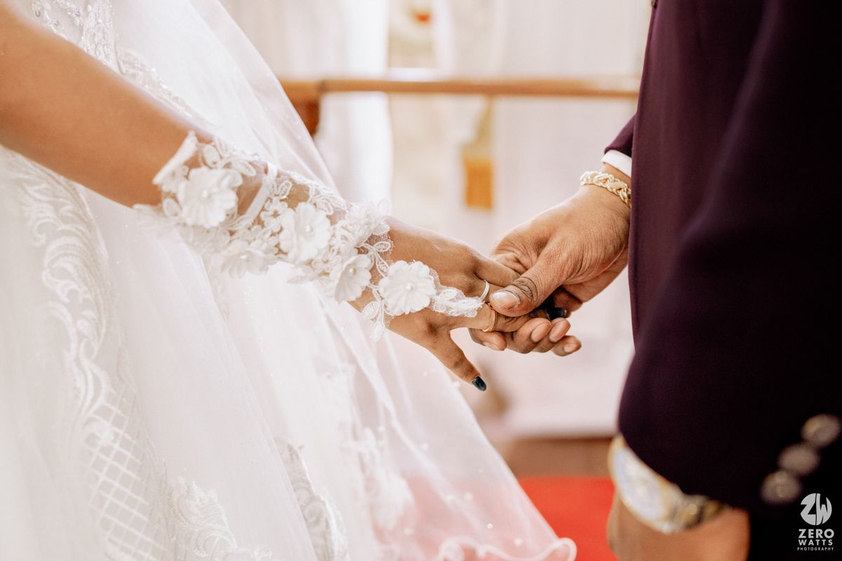Bride and groom holding hands during wedding ceremony, bride wearing lace gown with floral wrist detail.