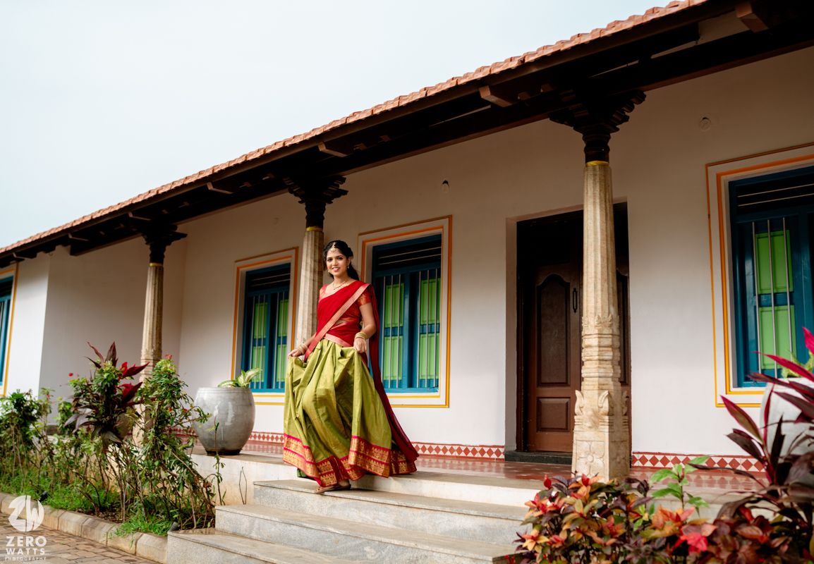 Bride in vibrant red and green silk saree walking outside a white traditional South Indian heritage bungalow.