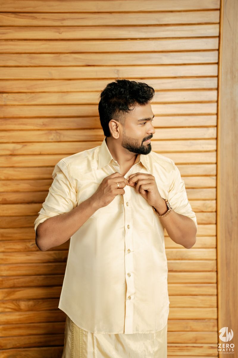 Man in cream silk shirt buttoning up against wooden slat background.