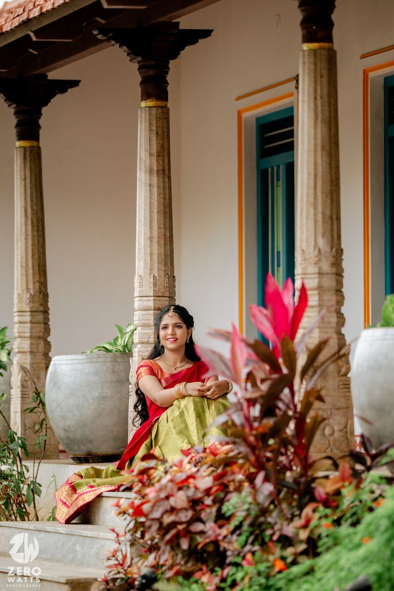 A bride in a red and green saree sits gracefully on temple steps surrounded by colorful plants and ornate stone pillars.