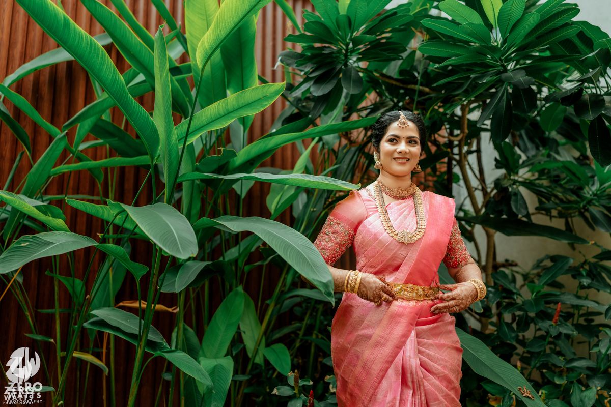 South Indian bride in pink silk saree with gold jewelry smiling outdoors among tropical green plants.