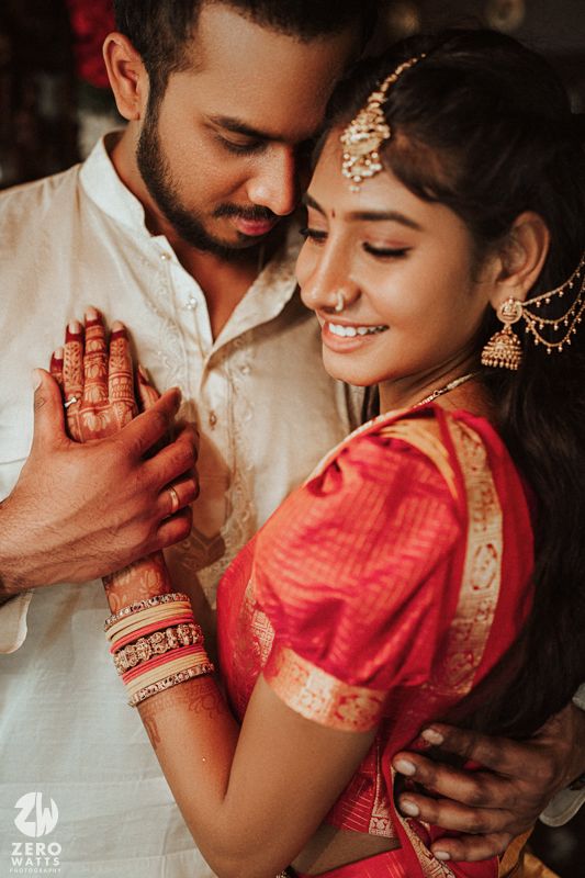 South Indian bride in red silk saree with gold jewelry sharing intimate moment with groom holding hands.